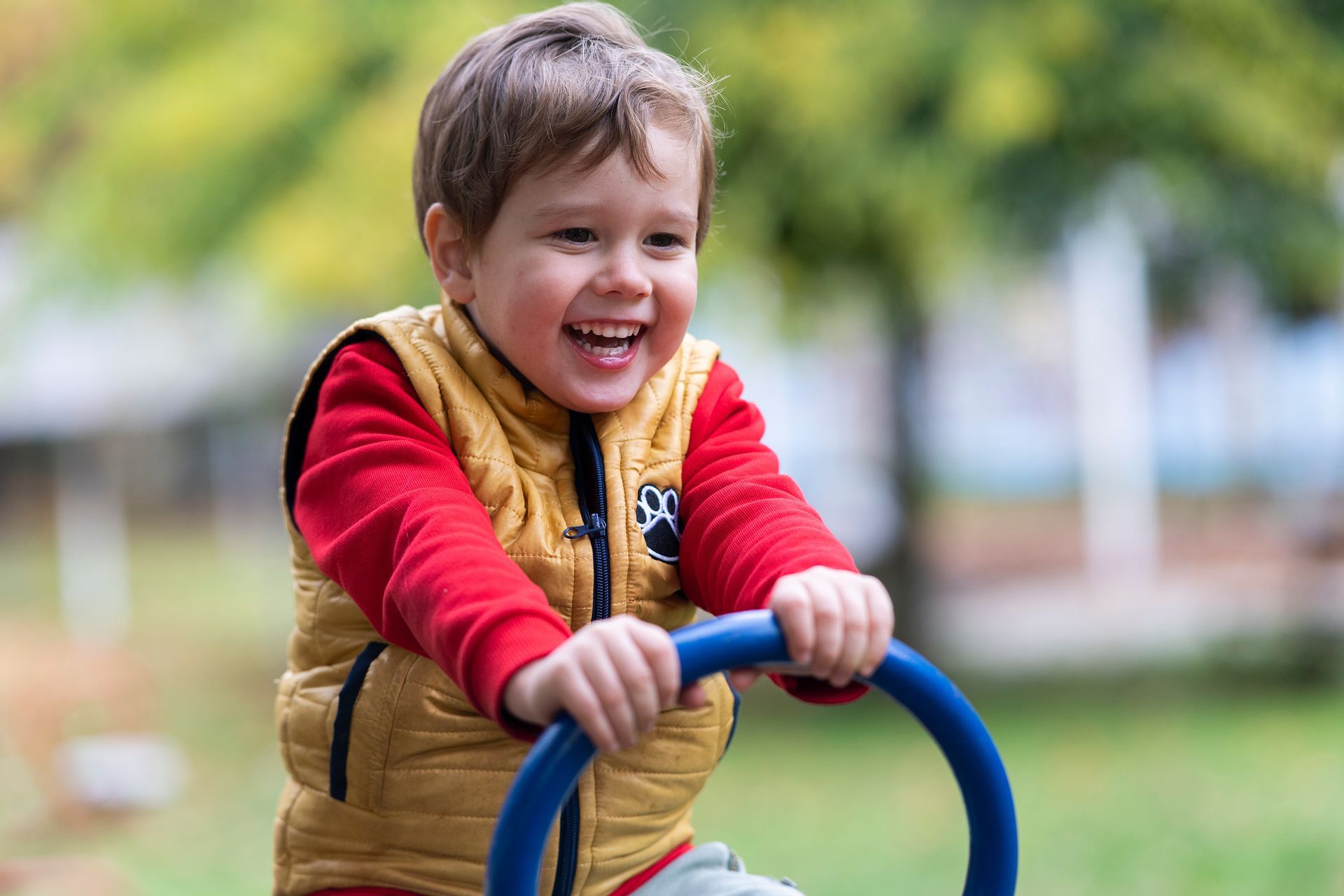 Boy smiling while holding a blue bar outdoors; wearing red shirt and yellow vest.