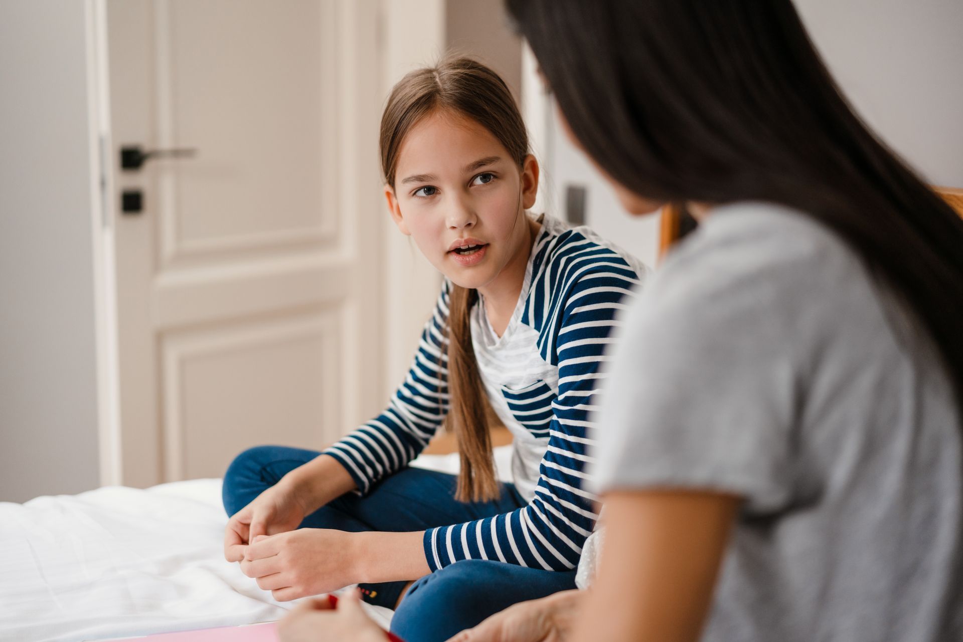 Girl in striped shirt talking to a person, both seated on a bed.