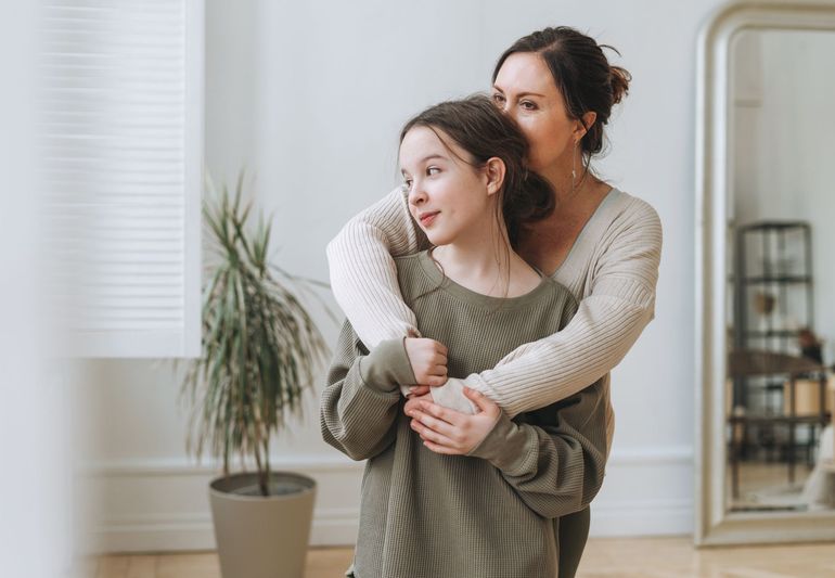 Woman hugging a young person indoors; both looking away with smiles.