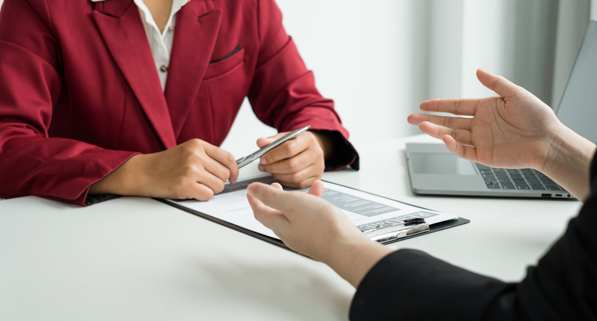 Two people at a desk: one in red blazer holding pen, other gesturing. Interview setting, white table.