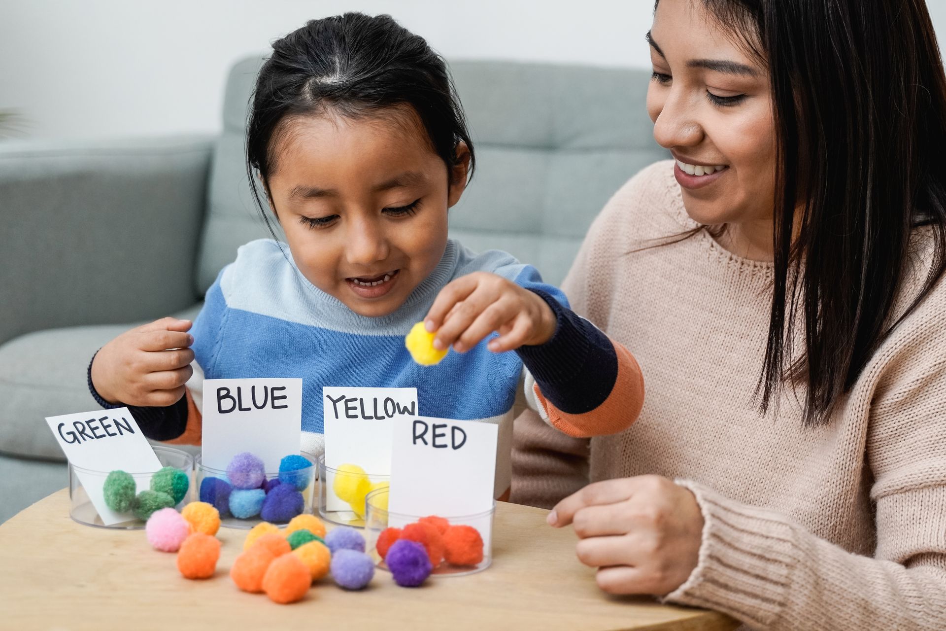 Child sorting colored pom-poms into labeled containers with an adult. They are smiling.