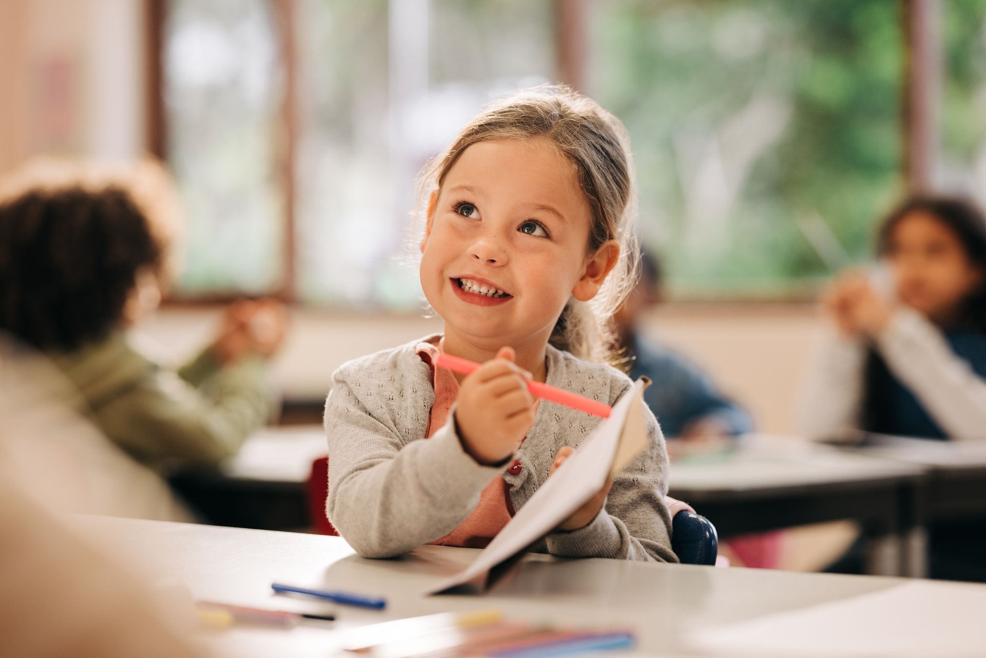 Smiling child in classroom, holding pencil and paper, looking up.