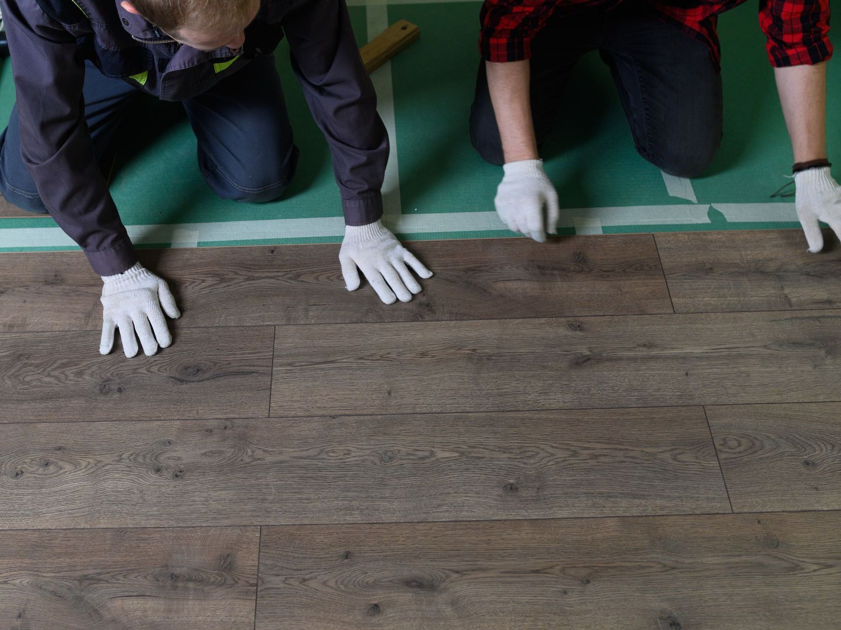 Three gloved workers kneel on a wooden floor, aligning planks beside a green taped edge.