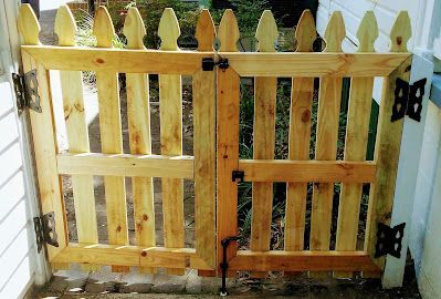 Wooden picket gate with vertical slats, ornate top, black hardware, attached to a white building.