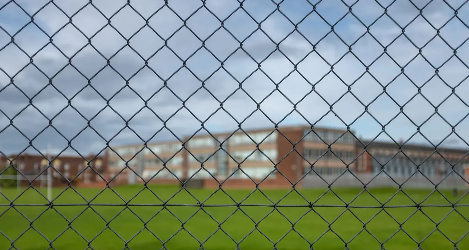 Chain-link fence with a rural backdrop of fields, buildings, and a lake under a blue sky.