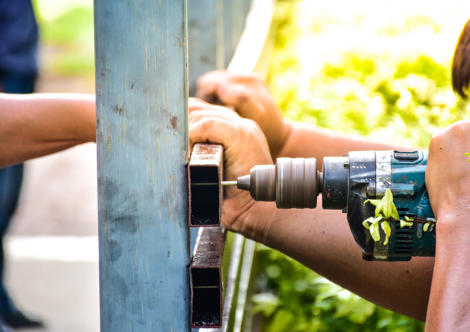 Person drilling a hole in a metal fence, with another person assisting outdoors in daylight.