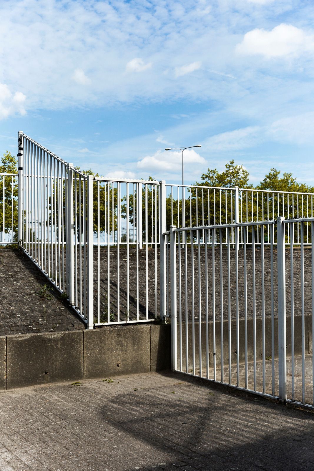White metal fence with open gate against a backdrop of trees and a blue sky.