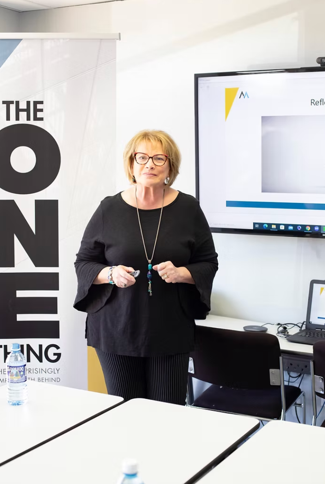 Woman standing in a classroom beside a screen and banner, holding a small device and smiling.