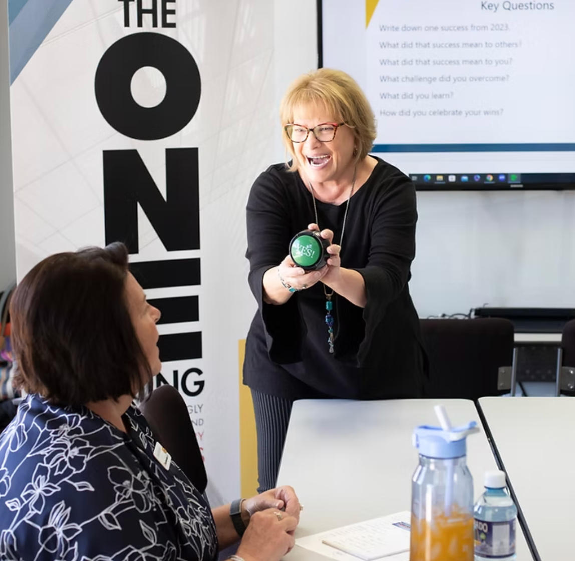 Sue woman in black demonstrates a green timer to a seated attendee in a bright meeting room.