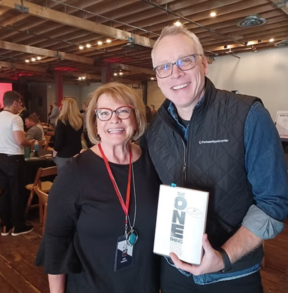 Two smiling people at an indoor event, holding a white “DINE” box.