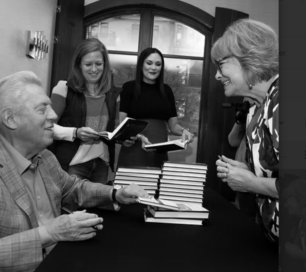 People gathering around a table with stacks of books, two women holding books and smiling.