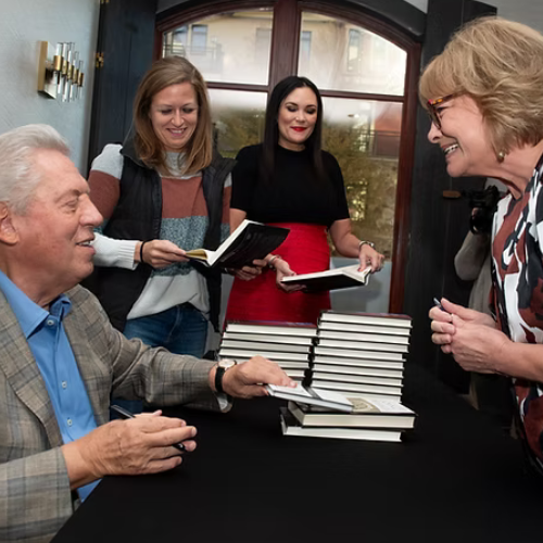 People gathered around a table with stacked books, smiling and talking in a cozy room.