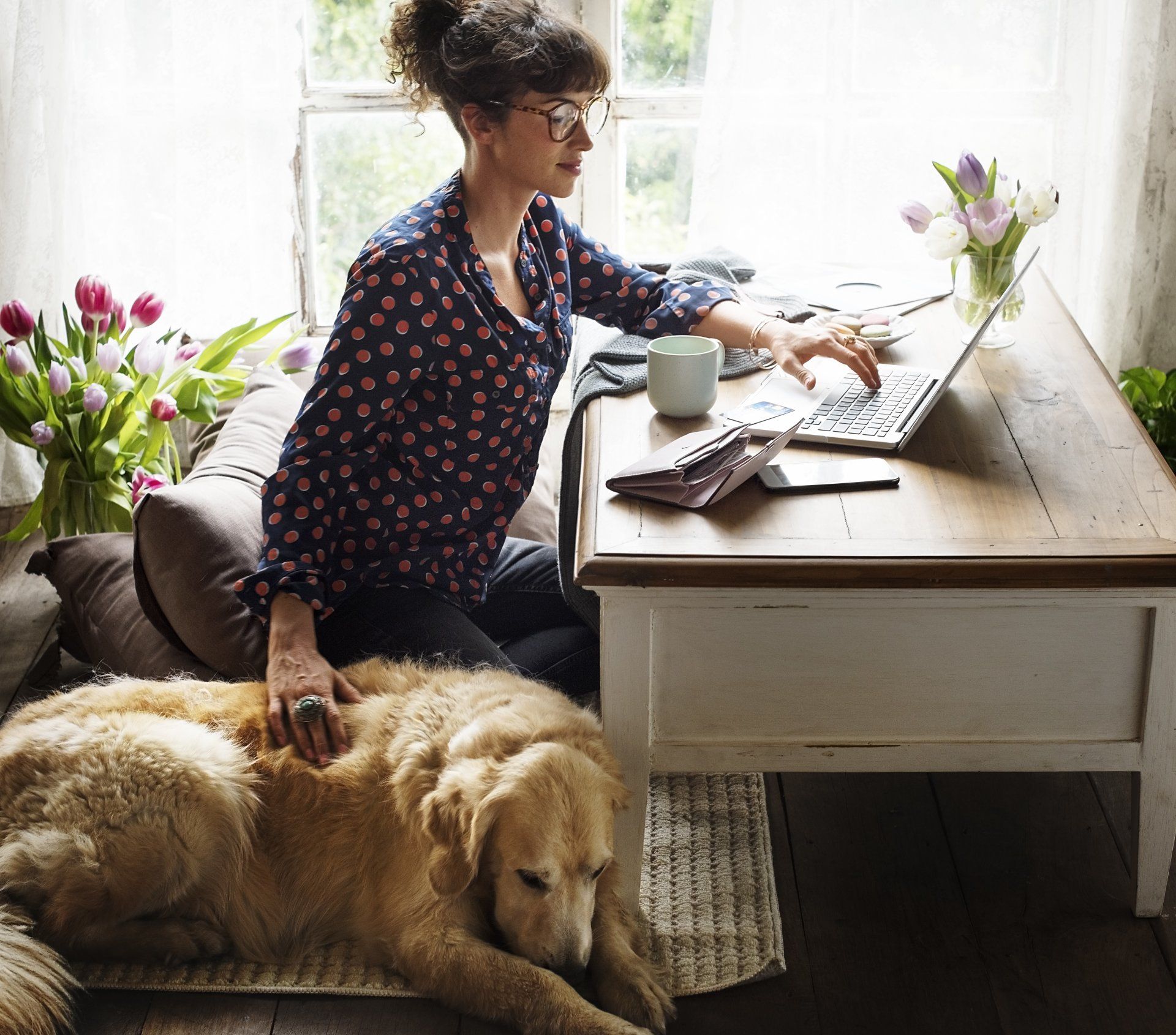 A woman is sitting at a desk with a laptop and a dog laying on the floor