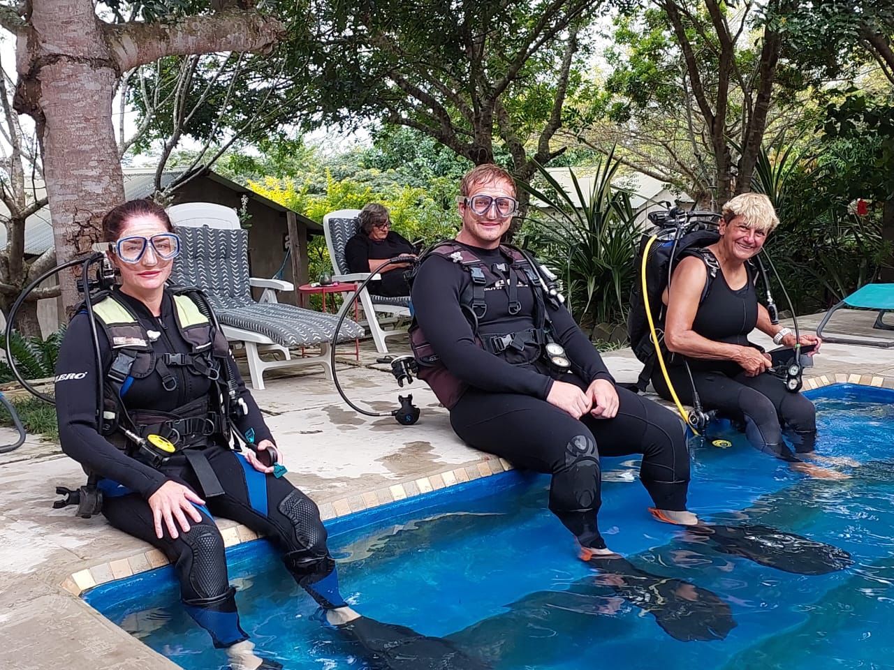 Three people in scuba gear sit at a pool edge, feet in the water, preparing to dive.