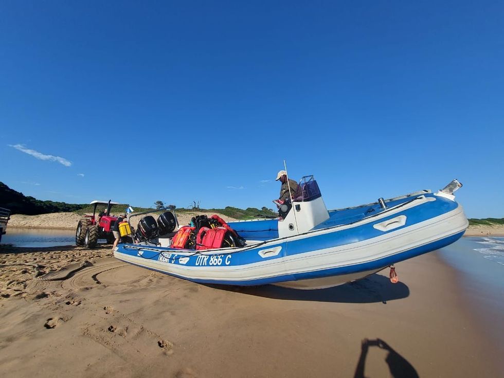 A blue and white inflatable boat on a sandy beach being pulled by a tractor under a blue sky.