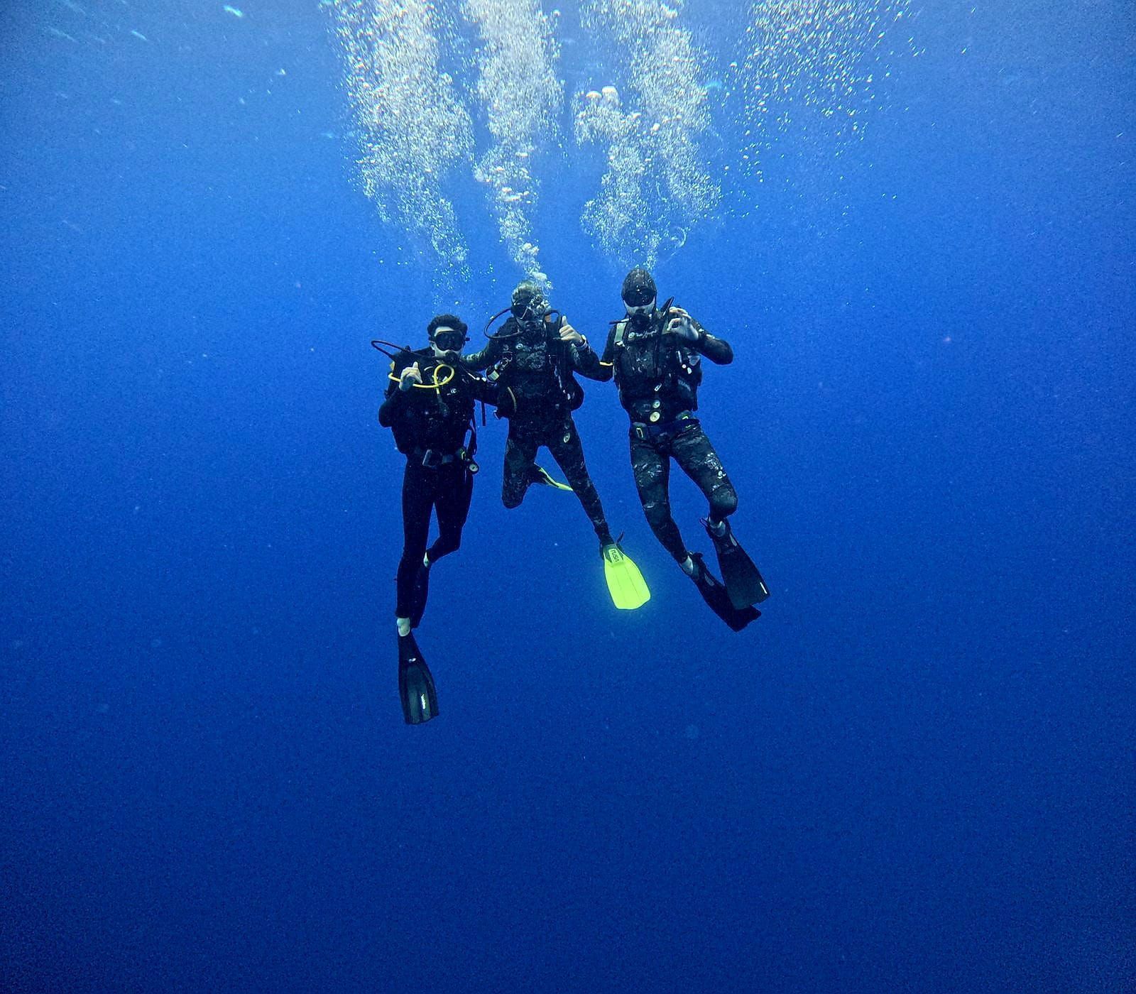 Three scuba divers swimming underwater, surrounded by blue water. Bubbles rise from the center diver's regulator.