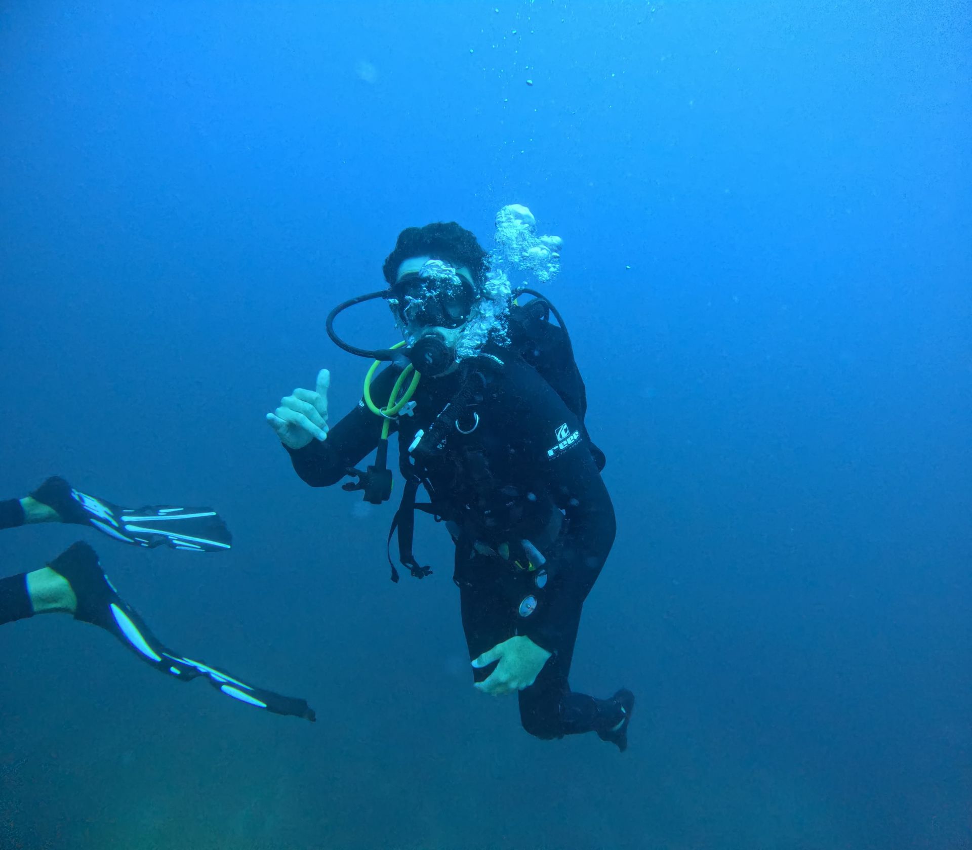 Scuba diver giving a thumbs-up underwater, surrounded by blue water. Black wetsuit, bubbles, flippers visible.
