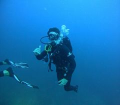 Scuba diver in black gear underwater, giving a thumbs-up, with another diver partly visible nearby.
