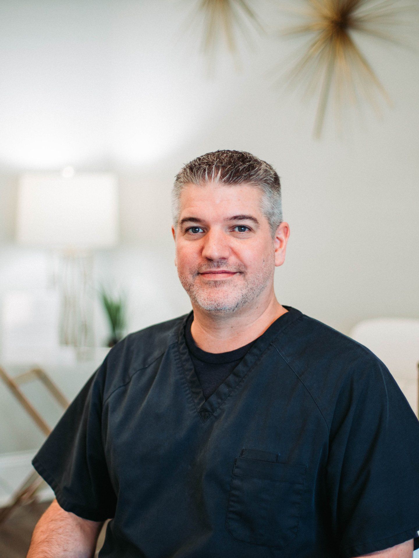 a man in a black scrub is sitting in a chair and smiling for the camera .