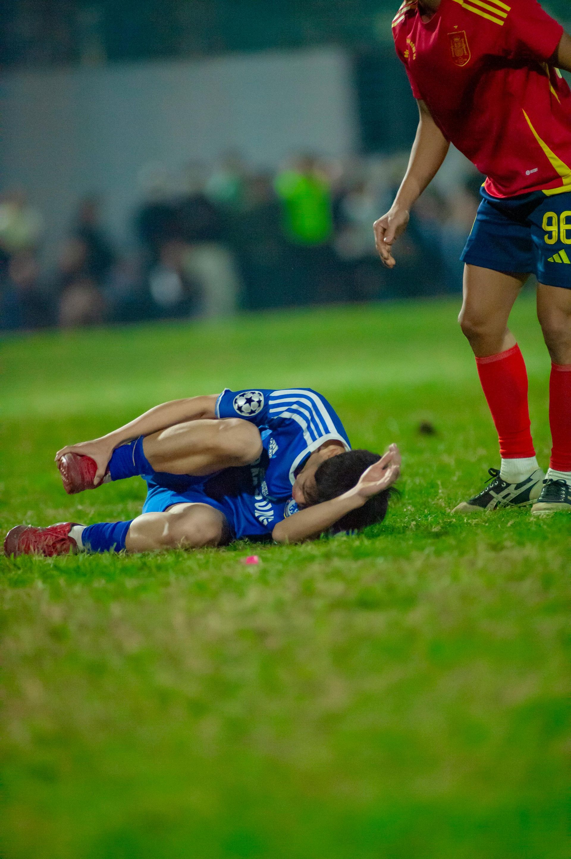 Blue soccer player lying on grass clutching his head as a red-uniformed opponent stands nearby