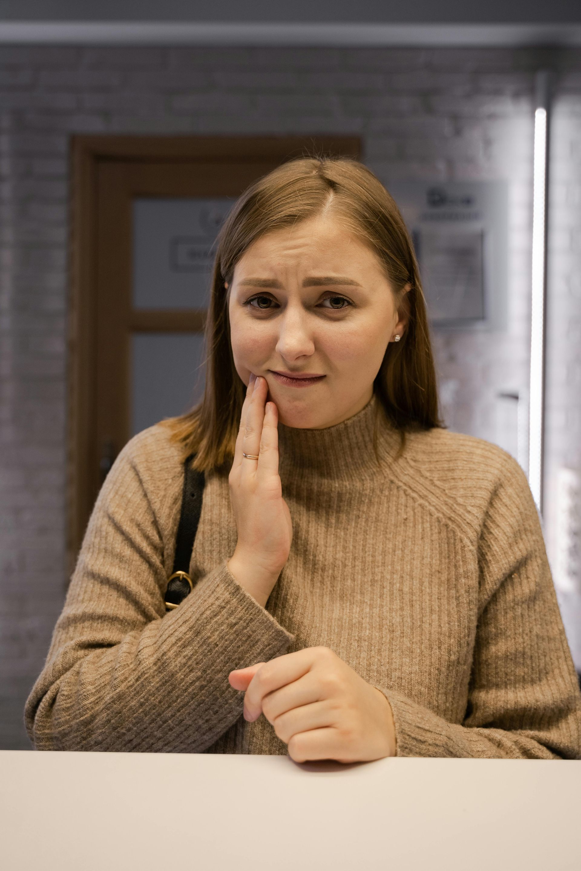 Woman in a beige sweater touching her cheek, standing indoors with painful expression.