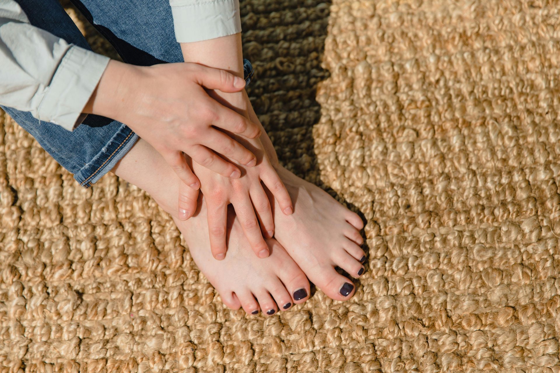 Two hands resting on bare feet with dark nail polish on a woven rug