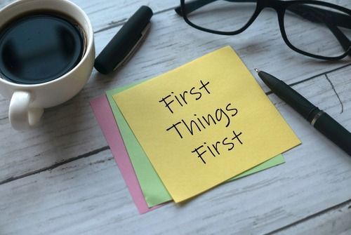 Sticky note reading “First Things First” beside coffee, glasses, and a pen on a desk