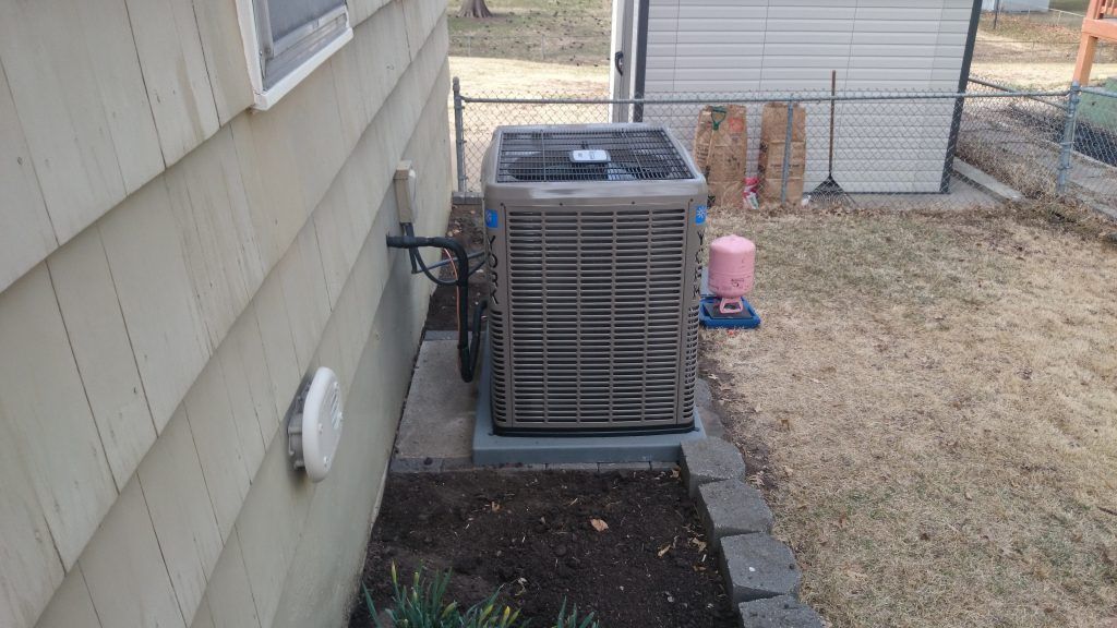 An air conditioning unit beside a beige house, located in a grassy yard. A fence and storage shed are in the background.