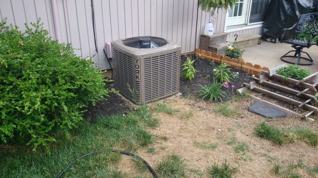 Air conditioning unit next to a house in a landscaped yard with bushes and a patio.