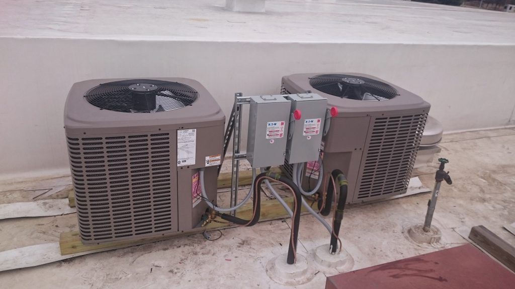 Two air conditioning units on a rooftop, connected by electrical boxes and piping. Grey and brown, sunny day.