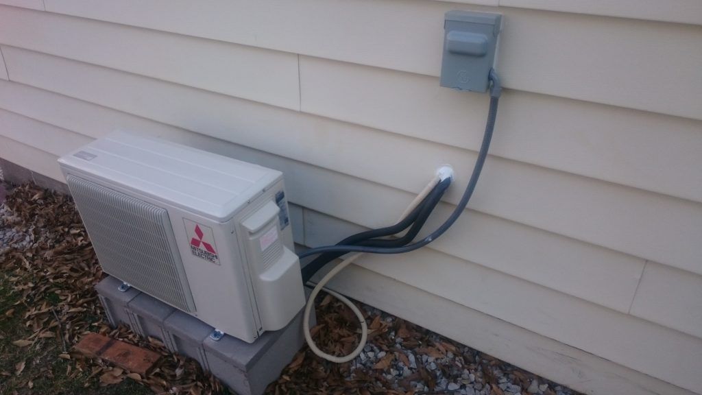 Mitsubishi air conditioning unit mounted on concrete blocks against a beige siding wall. Electrical components visible.