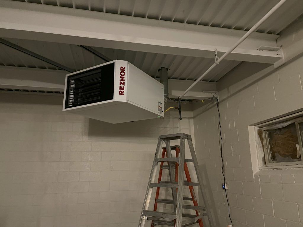 A white Reznor heater suspended from a white ceiling in an industrial setting, with a ladder below it.