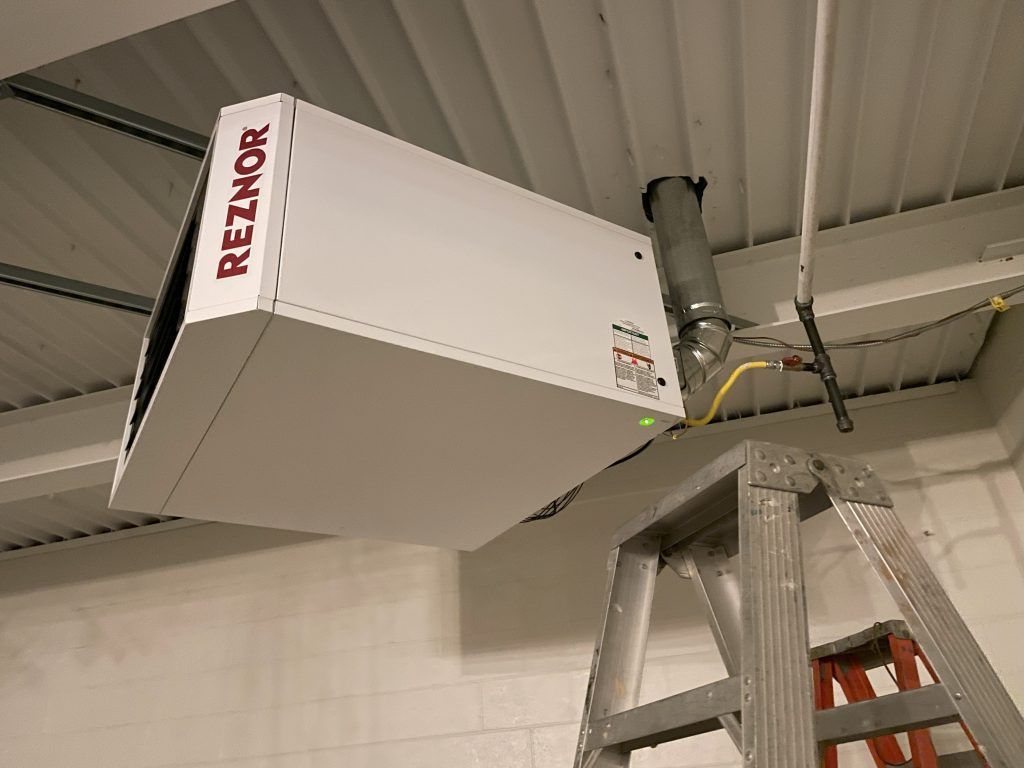 White Reznor heater mounted on a ceiling, with a ladder beneath it in an industrial building.