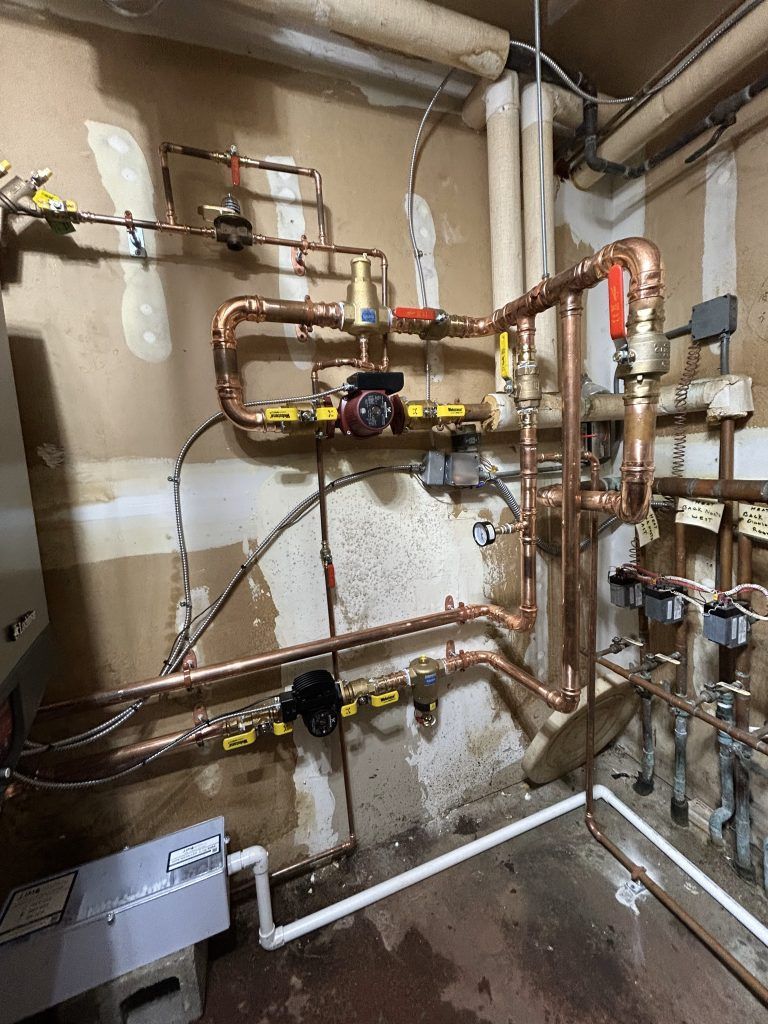 Copper pipes and valves on a wall, likely a utility room. Complex plumbing system with wiring and white wall.