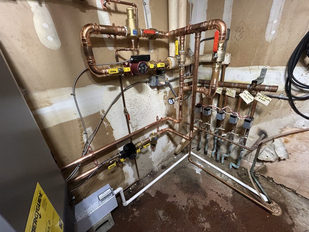 Copper pipes and valves of a heating system, on a wall with wiring and an attached pump.