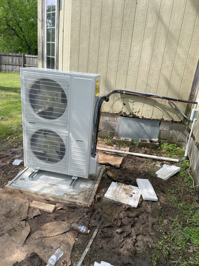 Gray air conditioning unit installed near a building on a concrete pad. Dirt and debris surround it.