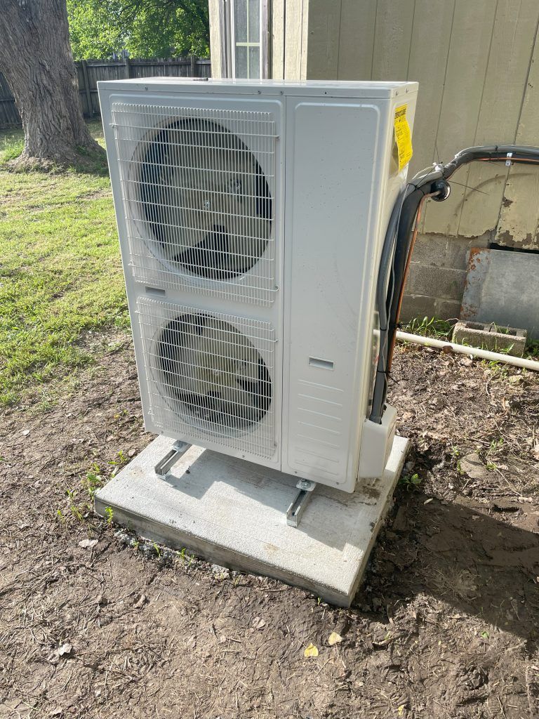 White HVAC unit mounted on a concrete pad outdoors, surrounded by dirt and a tree.