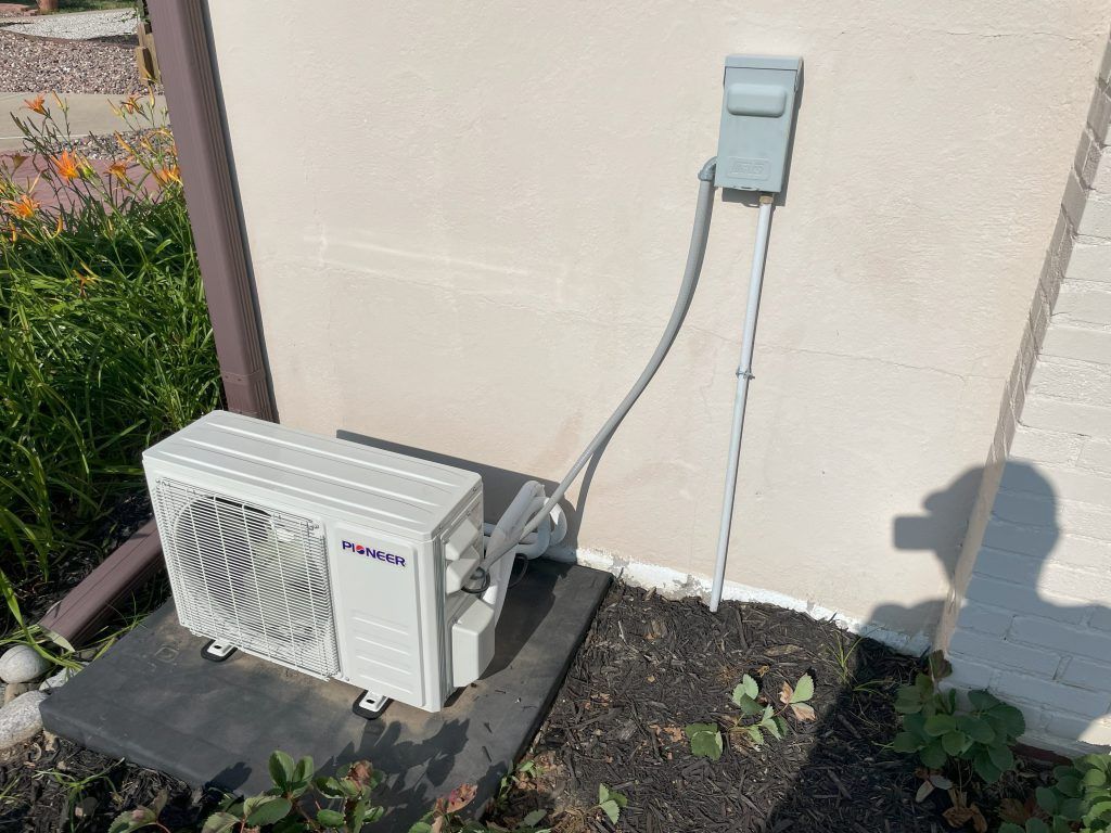 An outdoor air conditioner unit on a concrete base near a building. Gray conduit and electrical box are attached.