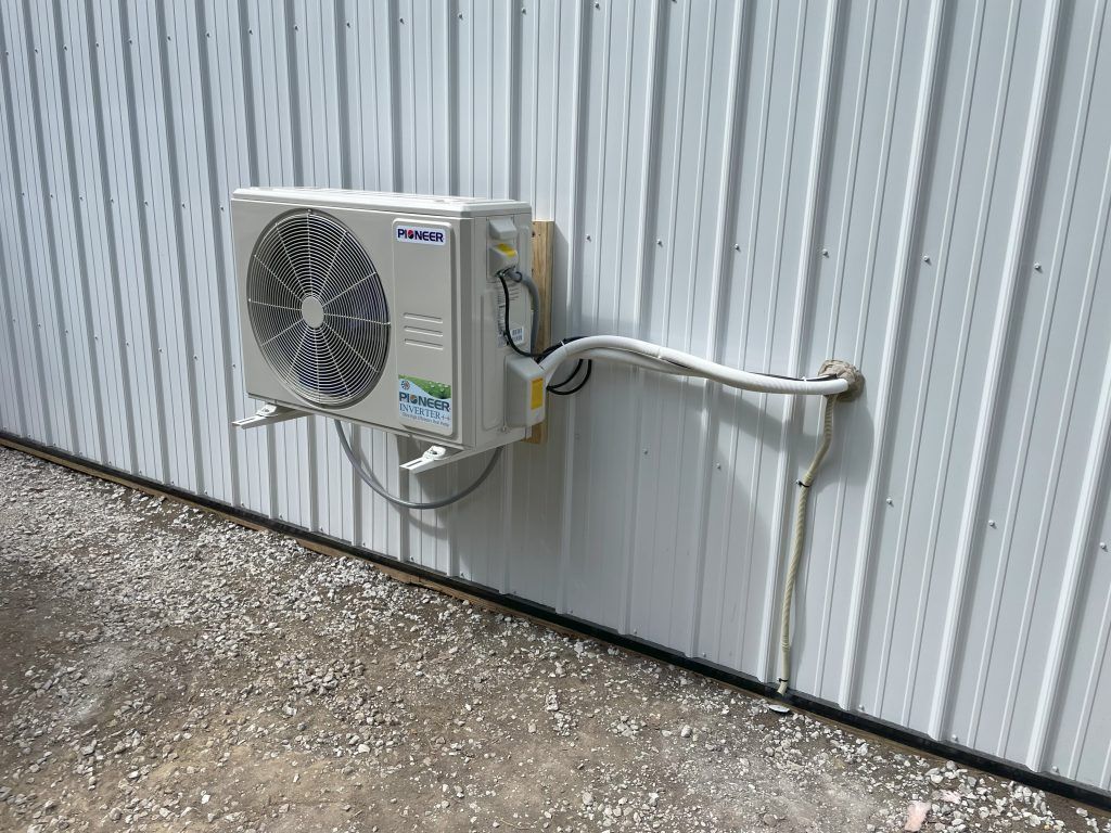 An outdoor air conditioning unit attached to a white corrugated metal wall with some ground gravel.