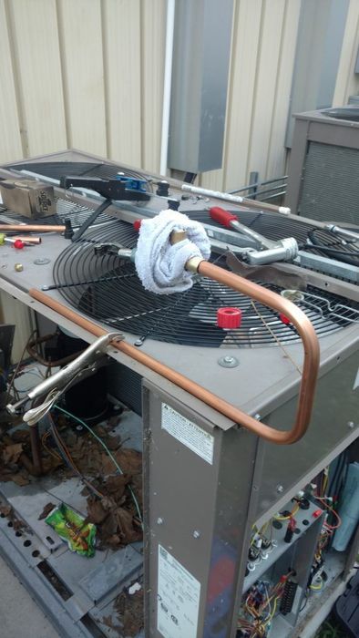 HVAC unit being repaired, with copper tubing, tools, and a wet rag covering a joint. Outdoor setting.