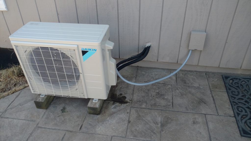 Daikin air conditioning unit on wooden blocks next to a wall, with water drain and electrical box.