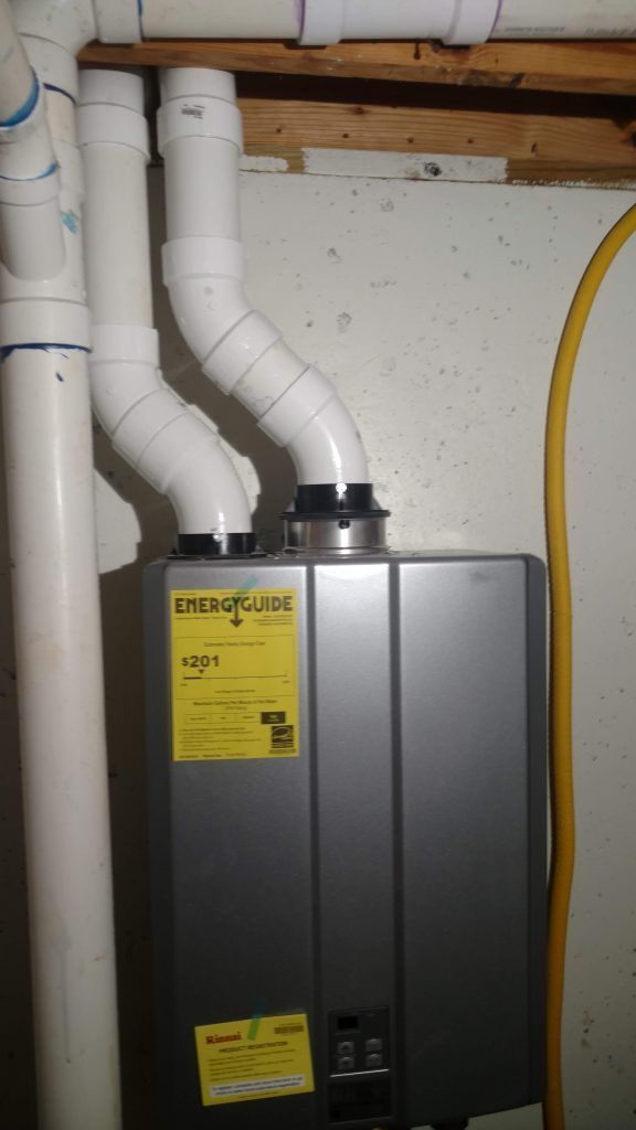 A tankless water heater with white PVC pipes venting upwards in a basement.