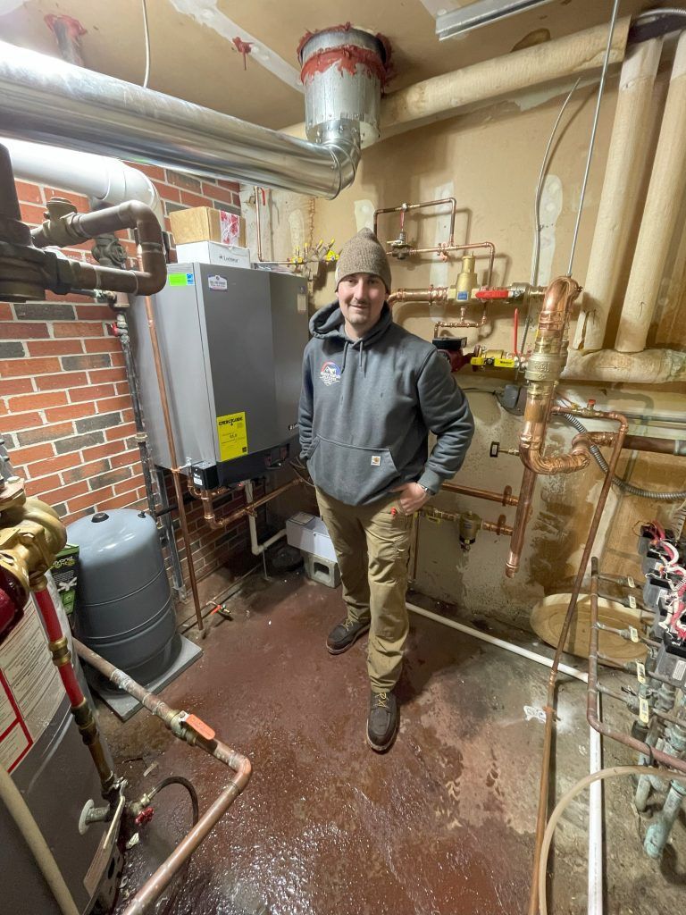 Plumber in a basement standing near a boiler and copper pipes, wearing a gray hoodie and brown pants.
