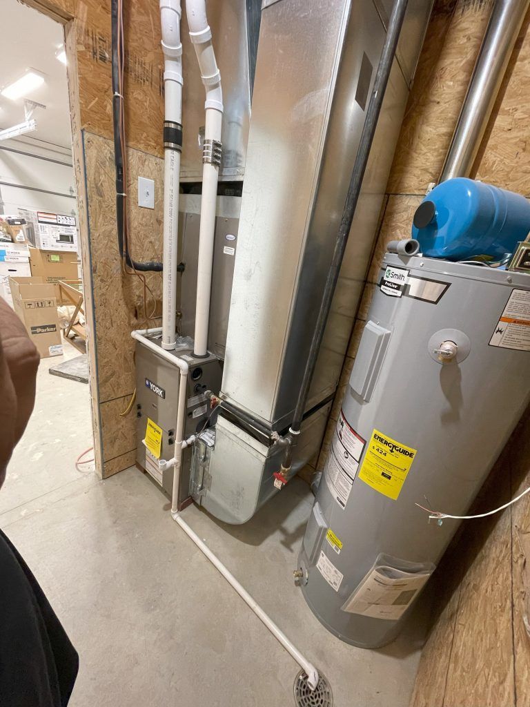 Furnace and water heater in a utility room. White pipes, gray metal, yellow stickers, and light brown wood.