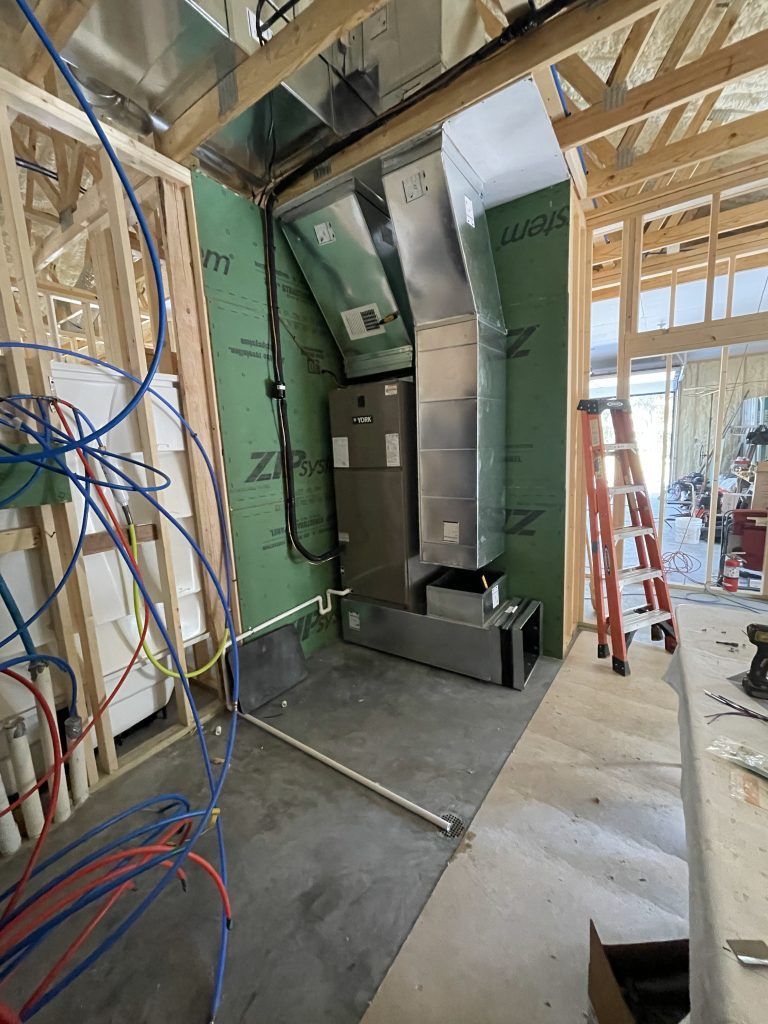 HVAC system installed inside a partially constructed building. Metal ductwork, green wall, and blue/red piping are visible.