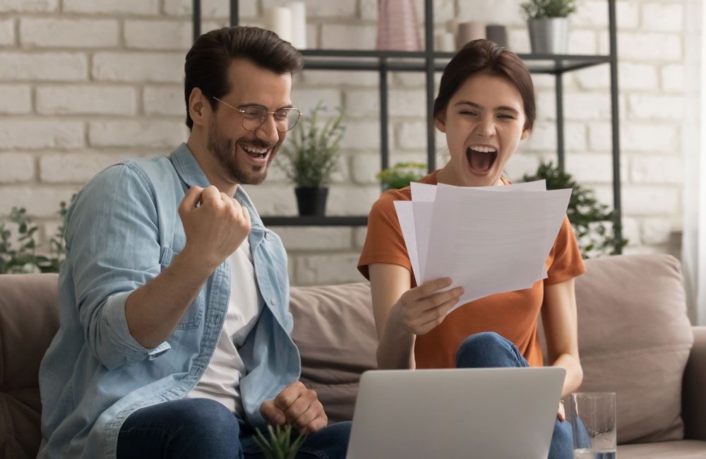 Couple excitedly celebrate, looking at papers, sitting on a couch near a laptop.