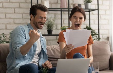 Couple excitedly celebrate, looking at papers, sitting on a couch near a laptop.