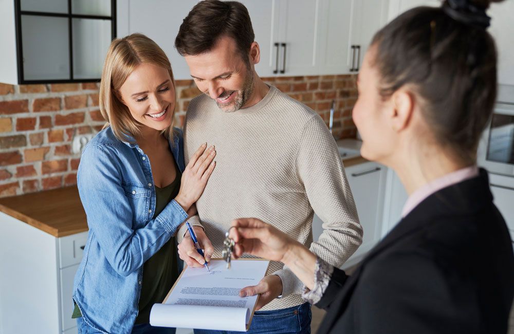 Couple signing document, receiving keys from real estate agent in kitchen.