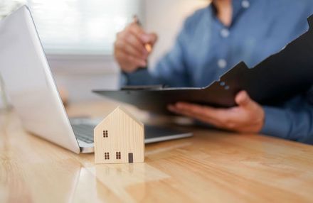Wooden house model on desk with laptop and person holding documents.