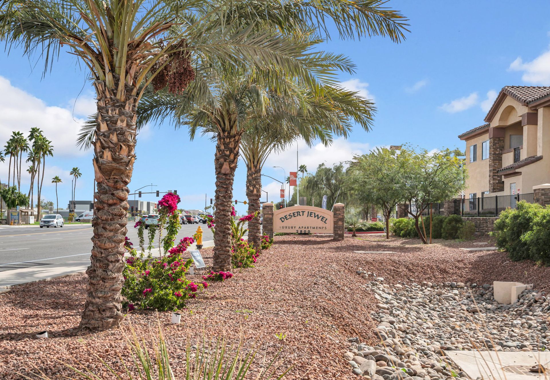 Entrance to a community with palm trees, a sign, and residential buildings under a blue sky.